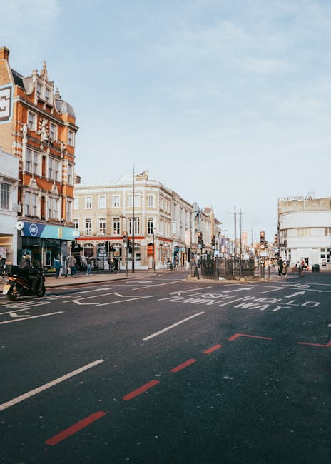 A wide view of a street scene in Swiss Cottage, featuring a mix of historic and modern buildings. On the left, there are multi-story brick buildings with ornate architectural details, including decora