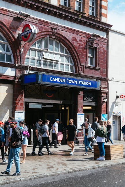 A wide view of a street scene in Swiss Cottage, featuring a mix of historic and modern buildings. On the left, there are multi-story brick buildings with ornate architectural details, including decora