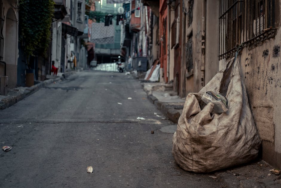 A large, weathered, off-white fabric sack filled with mixed waste materials, resting against a rough brick wall on a narrow urban street. The sack appears dirty and slightly sagging, with a transparen