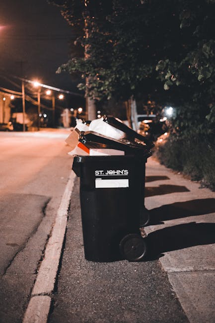 A large, weathered, off-white fabric sack filled with mixed waste materials, resting against a rough brick wall on a narrow urban street. The sack appears dirty and slightly sagging, with a transparen