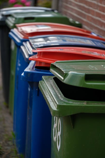 A large green waste disposal bin filled with an assortment of cardboard boxes and packaging materials in various sizes, some flattened and others partially assembled, with visible tape and labels. The