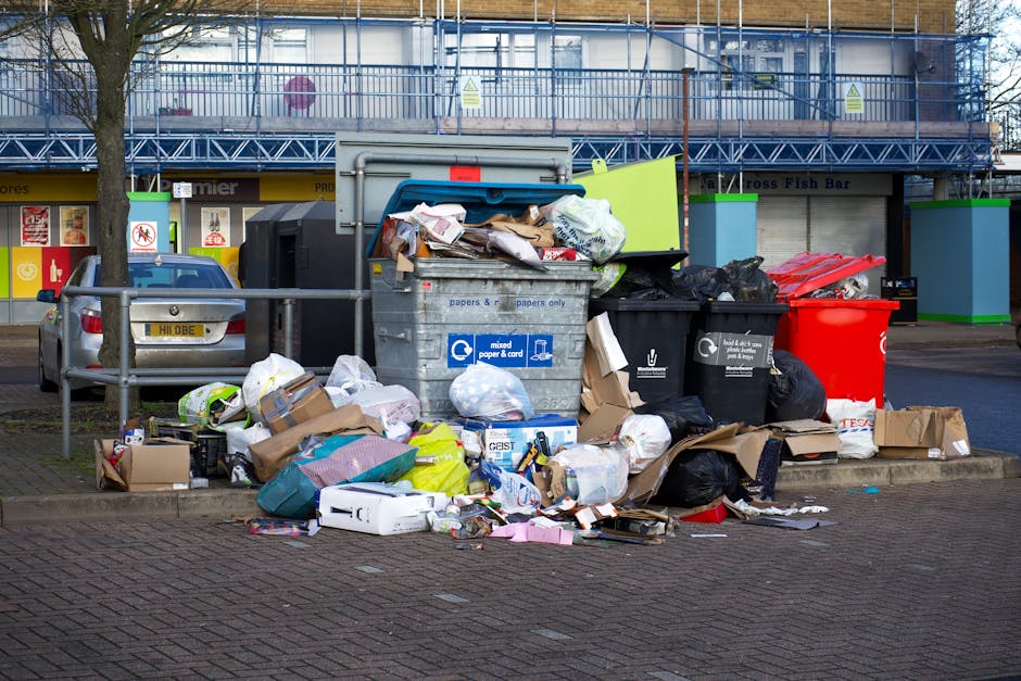 A large, green, weathered waste bin filled with various types of discarded packaging materials including cardboard boxes, paper forms, and plastic items, situated outdoors on a paved area. The bin is 
